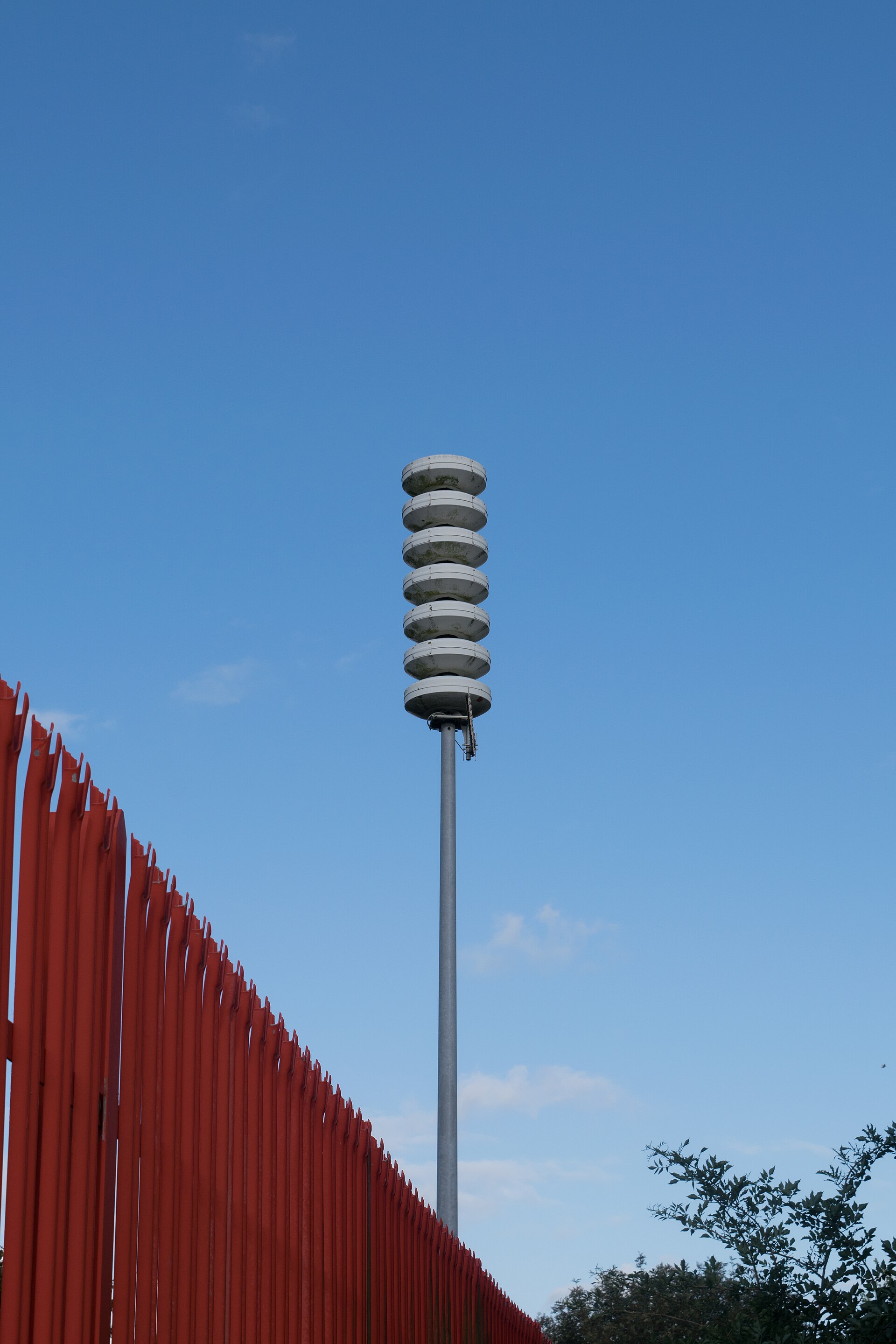 A stacked public warning siren on a tall mast against a clear blue sky, behind a red palisade fence.