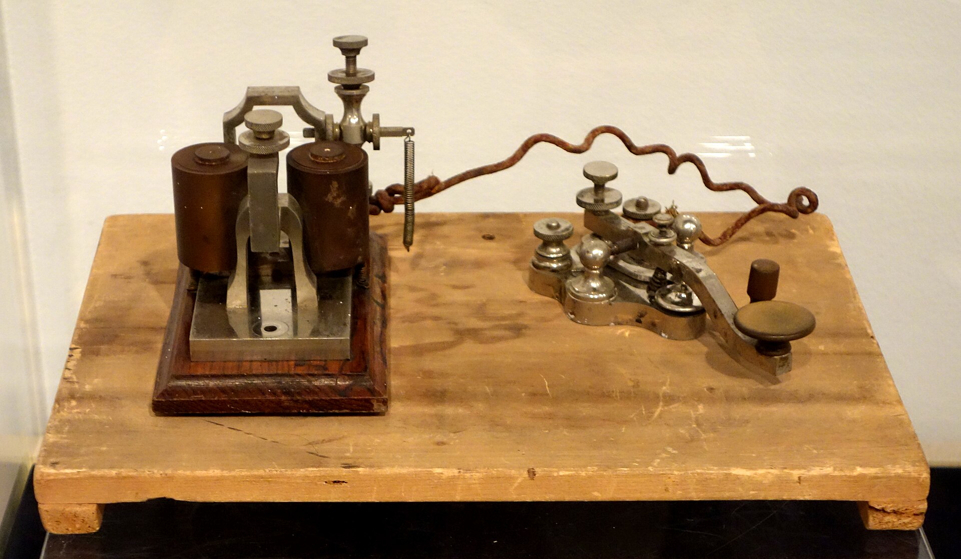 An 1876 telegraph key and sounder mounted on a pale wooden board, wired together with a coiled copper lead.